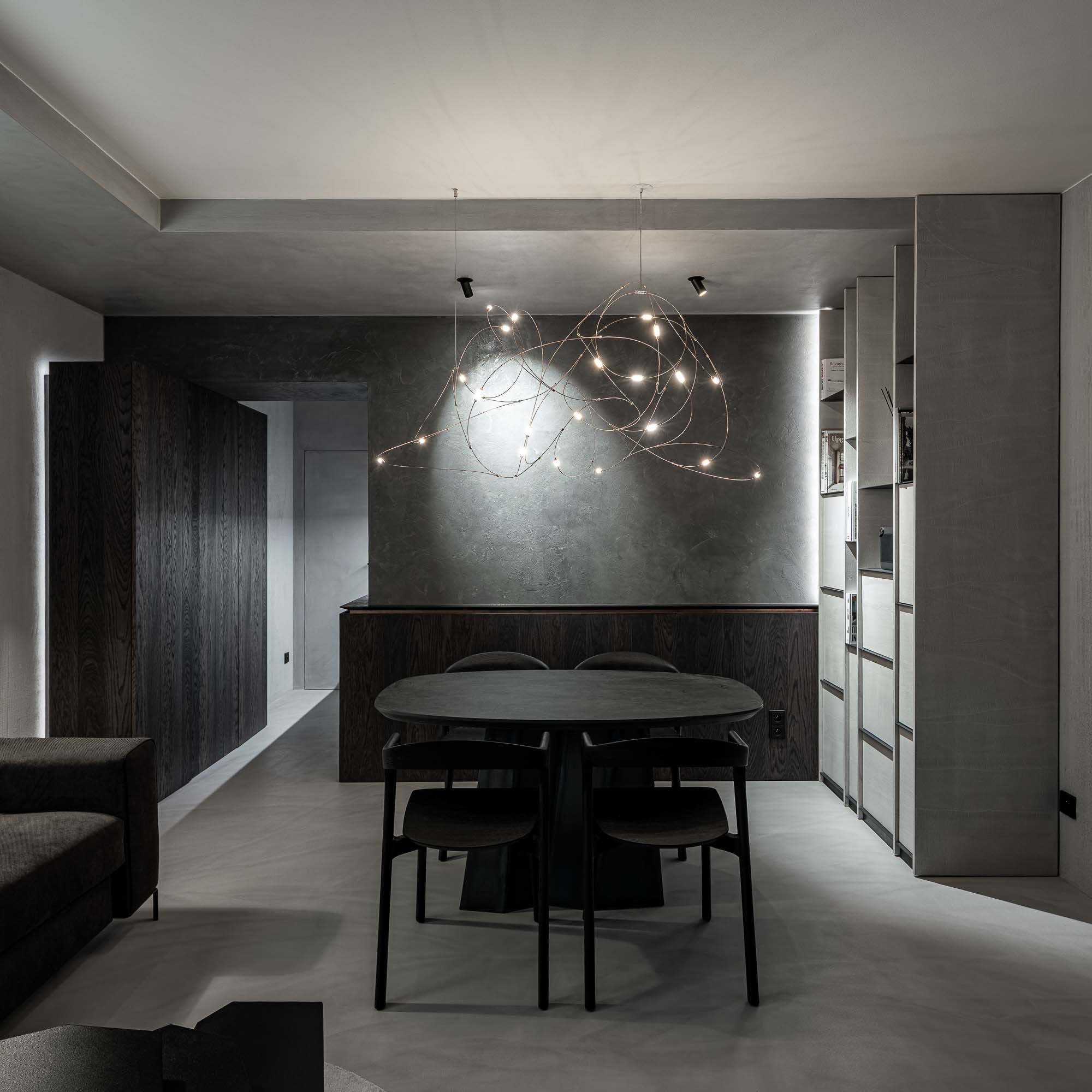 Central lounge space of Selection Properties — Ethnicraft Bok black oval table with four chairs, Moooi Flock of Light chandelier, dark plaster accent wall, built-in shelving on the right, kitchenette in the background on the left