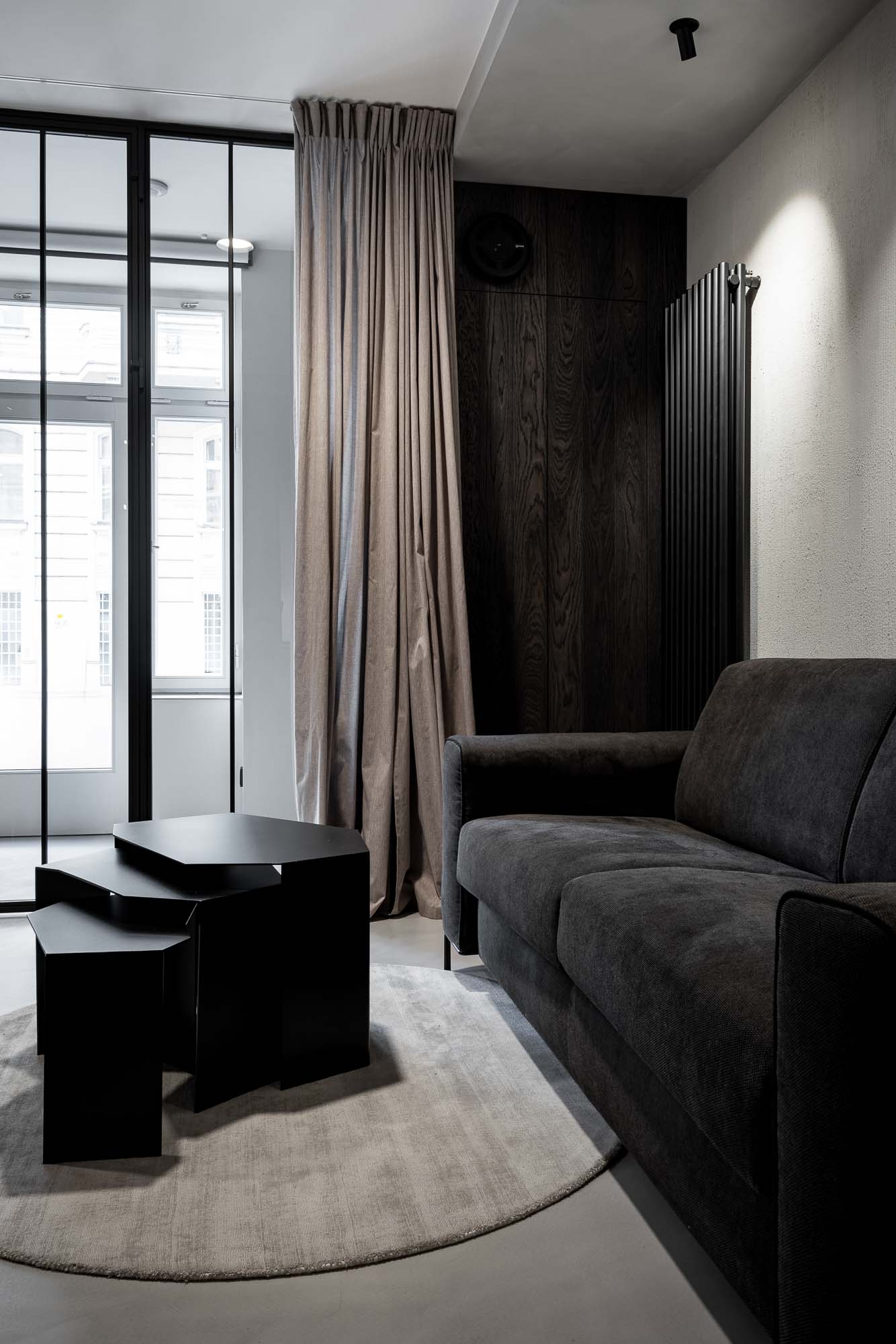Lounge zóna Selection Properties ve světle — tmavě šedá pohovka, asymetrické černé odkládací stolky, kulatý koberec, béžový závěs, ocelová prosklená příčka s denním světlem z ulice, černý vertikální radiátor vpravo