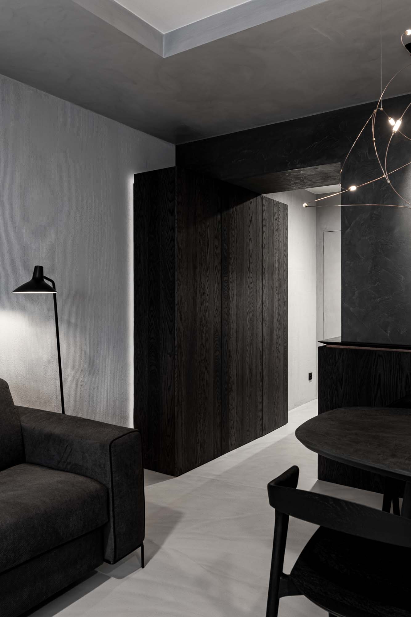 Pohled přes lounge k jídelní zóně Selection Properties — tmavý objem vestavné skříně z dřevěné dýhy, stojací lampička, pohovka vlevo, stůl Ethnicraft Bok vpravo, lustr Moooi Flock of Light v pozadí