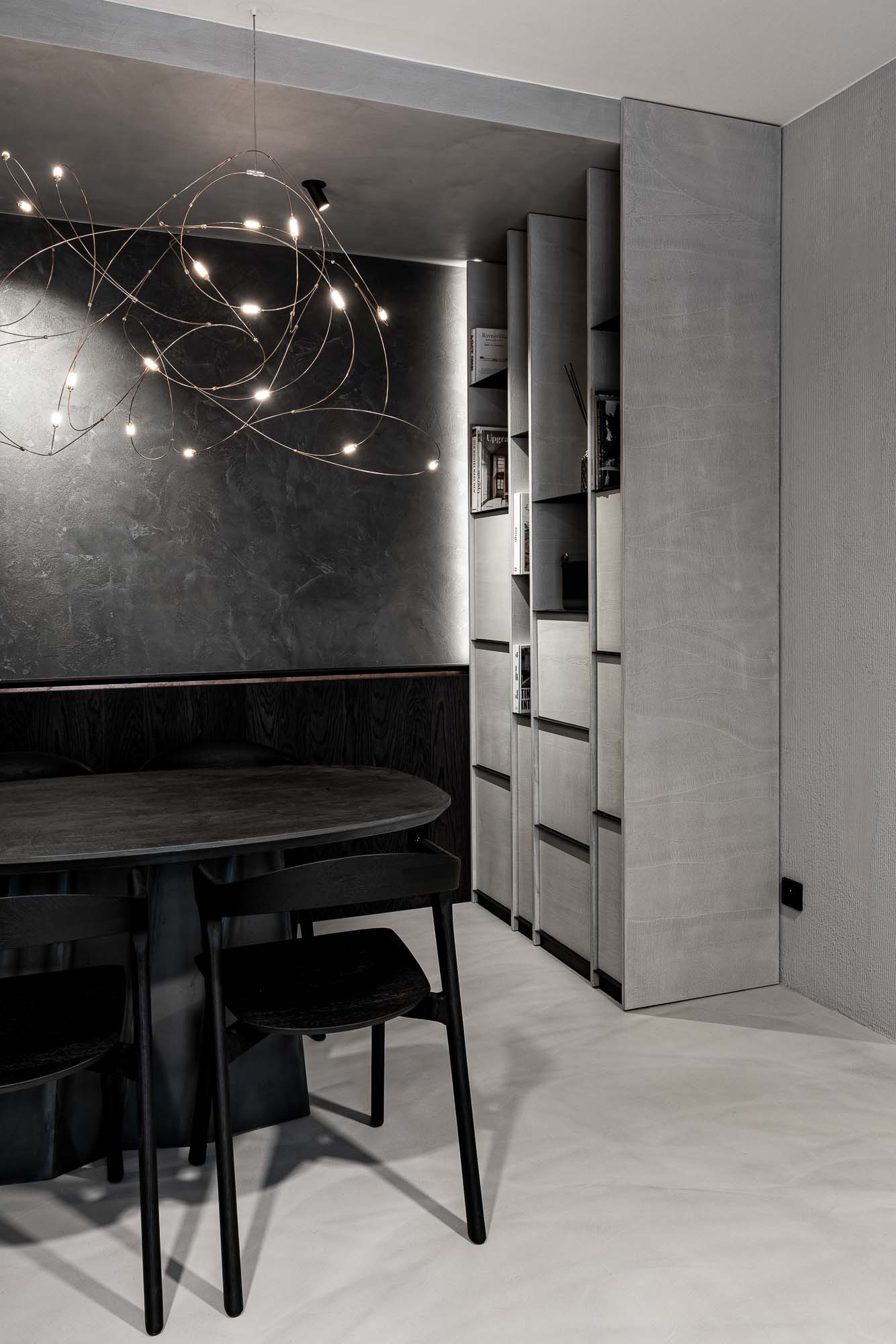 Pohled na kuchyňku a knižní stěnu Selection Properties — lustr Moooi Flock of Light vlevo, jídelní stůl s křesly, vestavná knihovna vpravo, skrytá kuchyňka za tmavým kuchyňským pultíkem