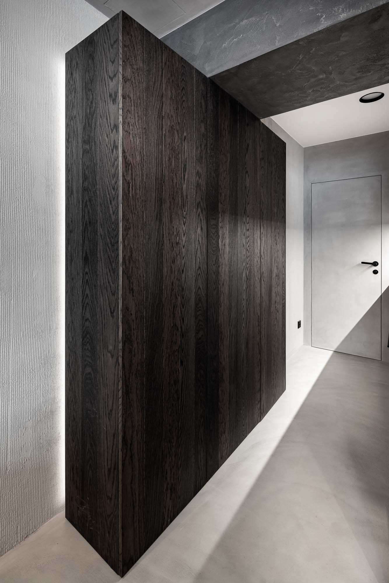 Chodba Selection Properties — tmavý objem vestavné skříně z dřevěné dýhy s LED podsvícením při podlaze, bezobložkové stěrkové dveře, cementová stěrka na stěnách a stropě