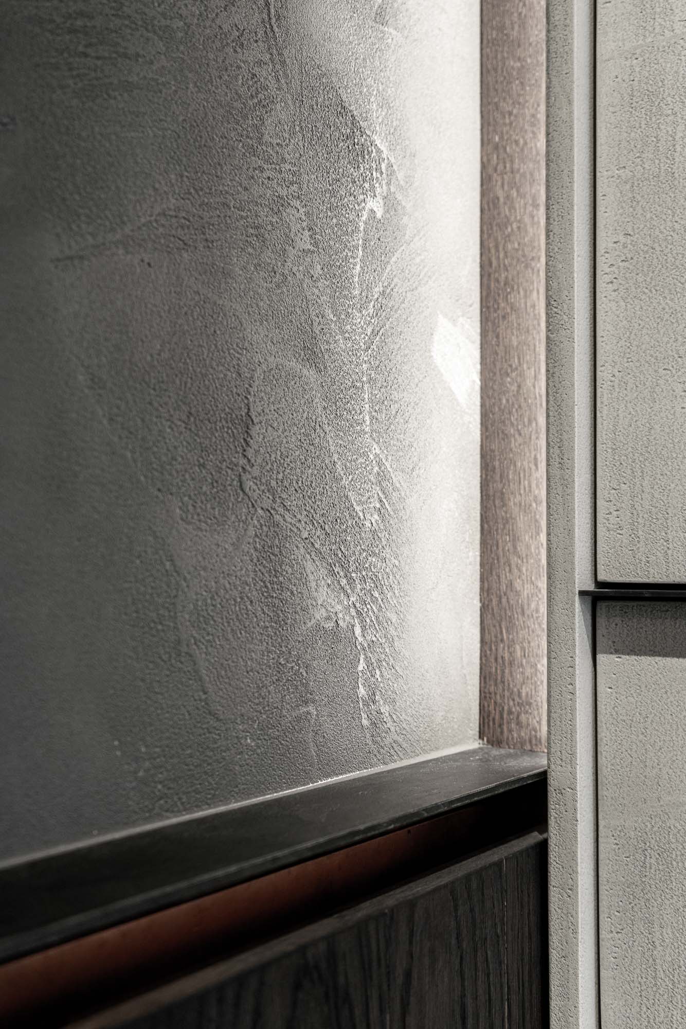 Detail přechodu cementové stěrky a dřevěné dýhy Selection Properties — světlá stěrka setkává s tmavě mořenou dřevěnou dýhou, měděná liniová hrana jako dekorativní prvek