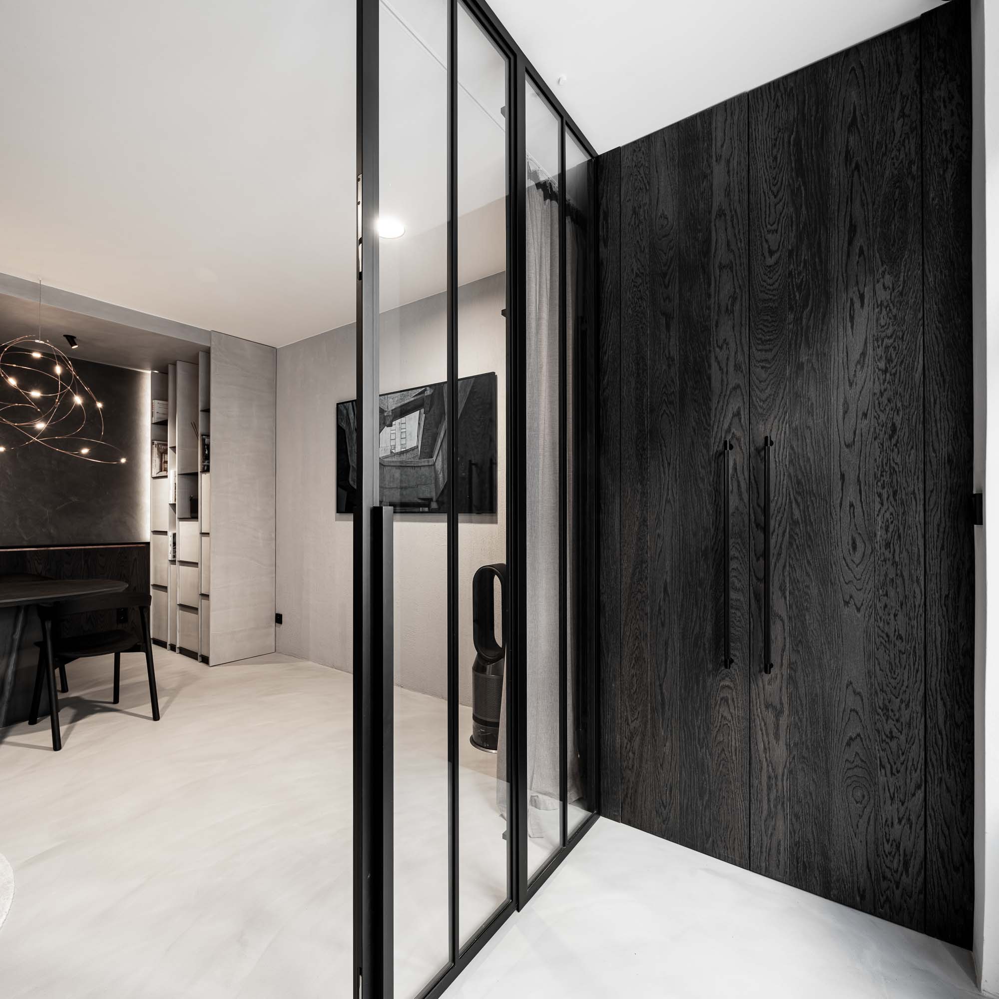 Pohled přes prosklenou příčku do lounge Selection Properties — ocelová prosklená příčka s Crittal profilem, tmavě mořená dřevěná skříňová stěna, lustr Moooi Flock of Light v dálce, cementová stěrka