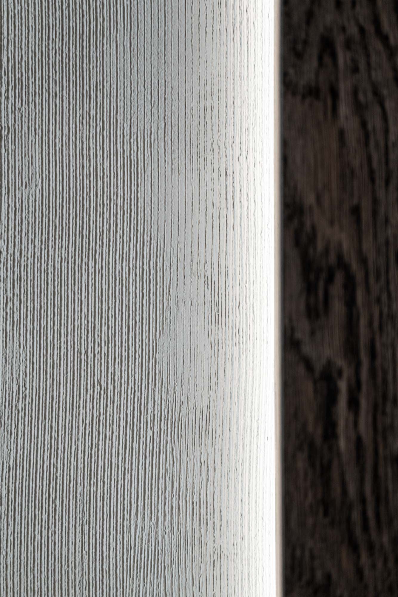 Detail přechodu materiálů Selection Properties — světlá cementová stěrka setkává s tmavou dřevěnou dýhou, podsvícená hrana odhaluje přesnost detailu